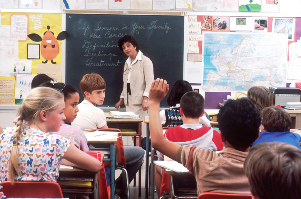an image depicting children in a classroom, with a child raising his left arm to ask the teacher a question