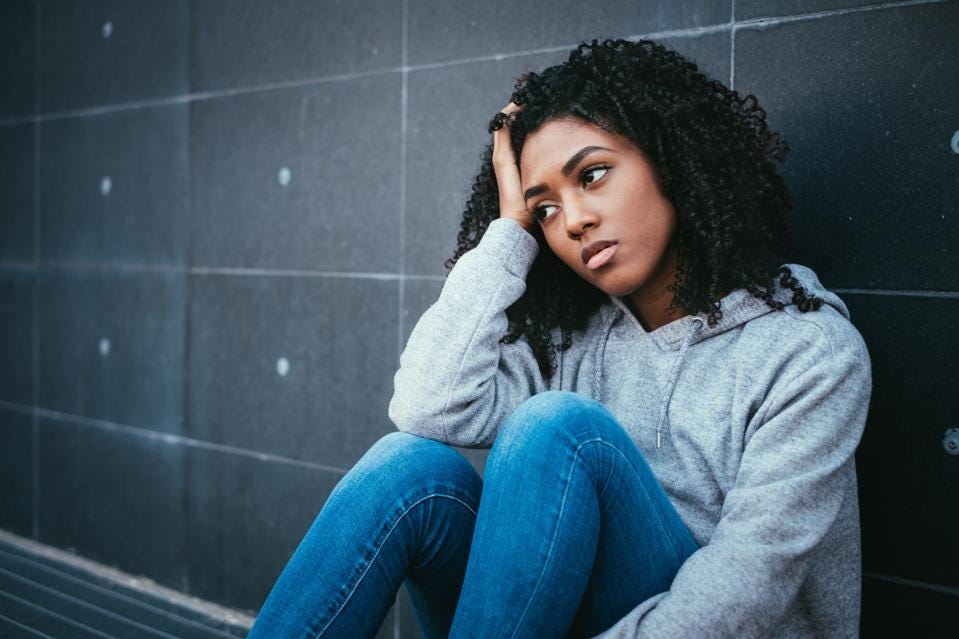 An image of a teen looking worried while sitting against a wall, with her right hand arm on her knee, supporting her head